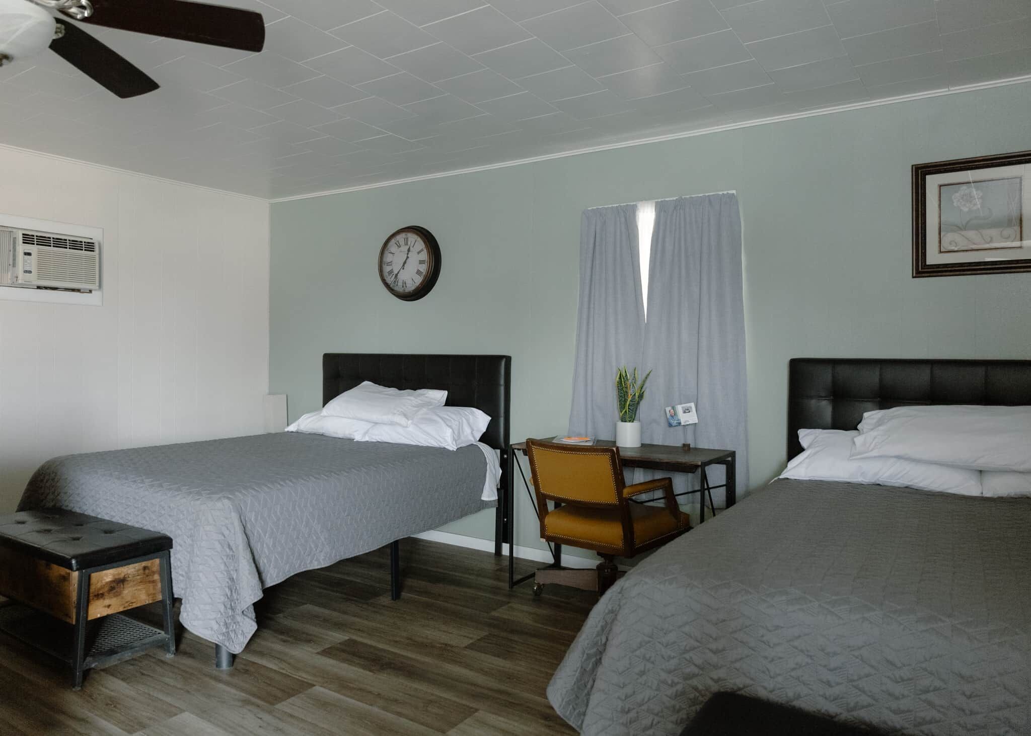 Een hotelkamer met twee bedden, een bureau, een stoel, een klok en een raam met gordijnen.