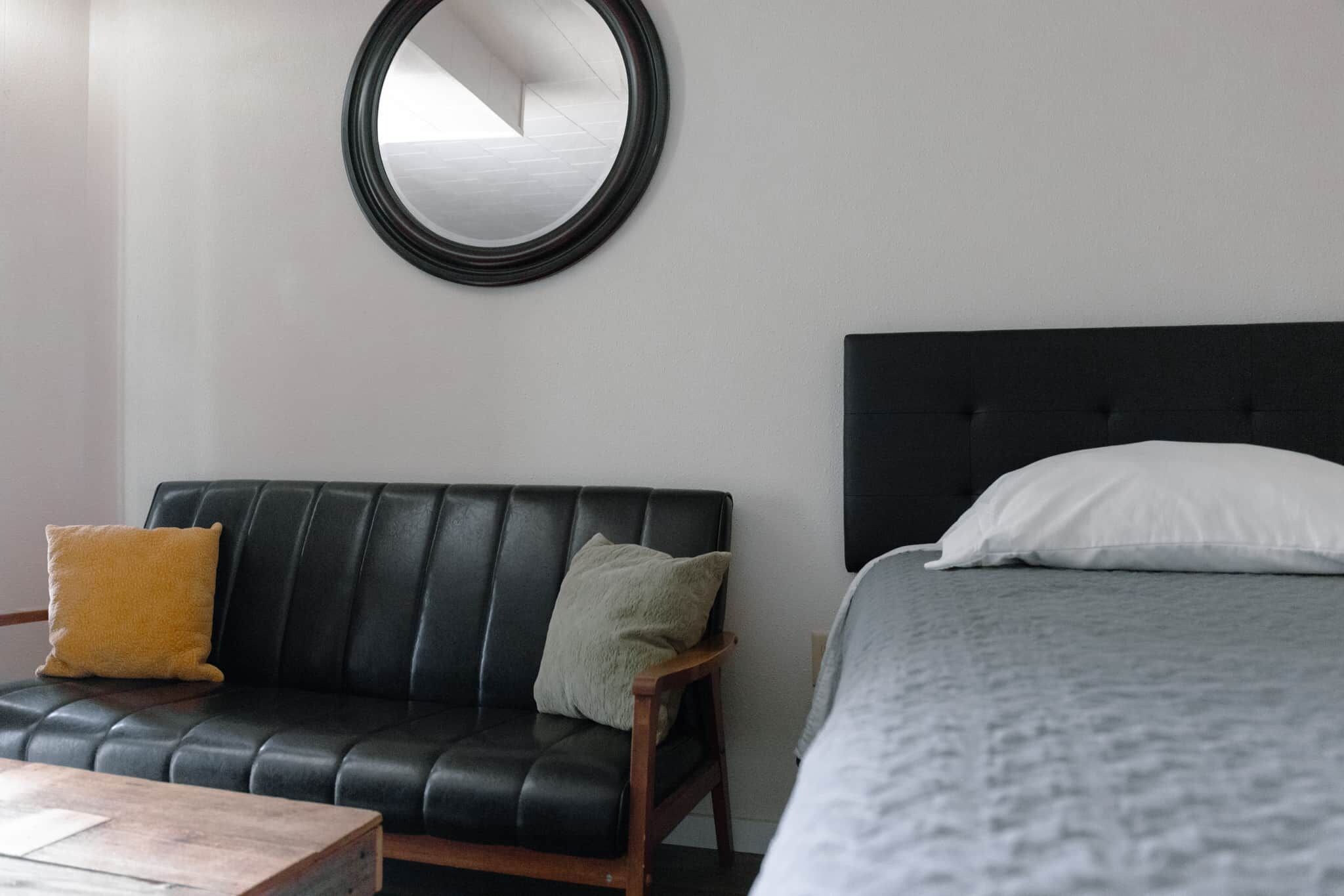 Una habitación con sofá de cuero negro, una silla de madera, cama con almohada blanca y un espejo redondo en la pared.