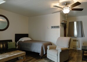 Un dormitorio con cama, una silla, un banco de madera, un ventilador de techo y una ventana con cortinas.