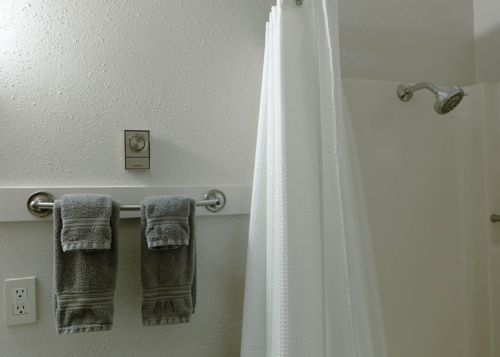 A bathroom with a towel rack holding two towels, a showerhead, and a thermostat on the wall.