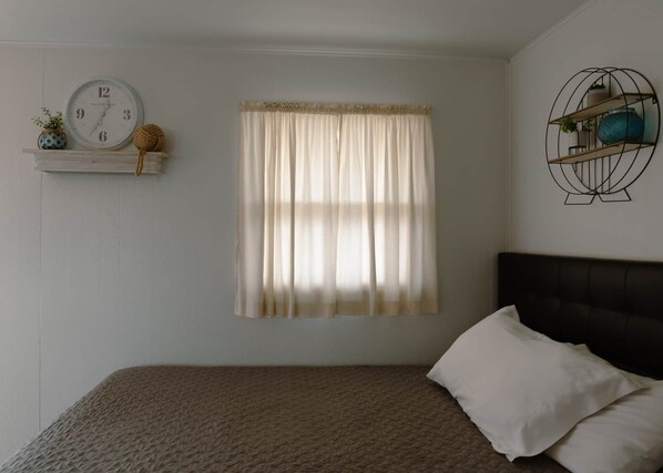 Quarto com cama, janela com cortinas, relógio na parede e prateleira com objetos decorativos.
