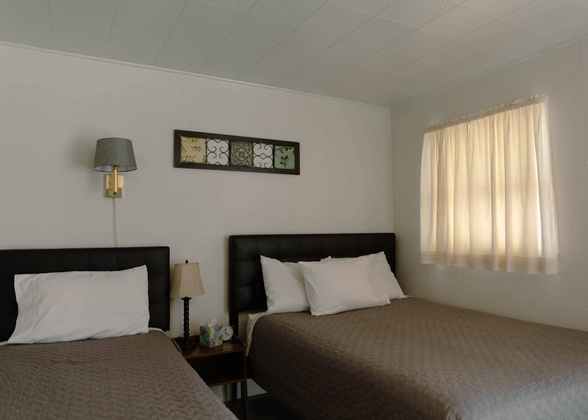 Une chambre d’hôtel avec deux lits, une table de chevet avec une lampe, une horloge et une fenêtre avec des rideaux.