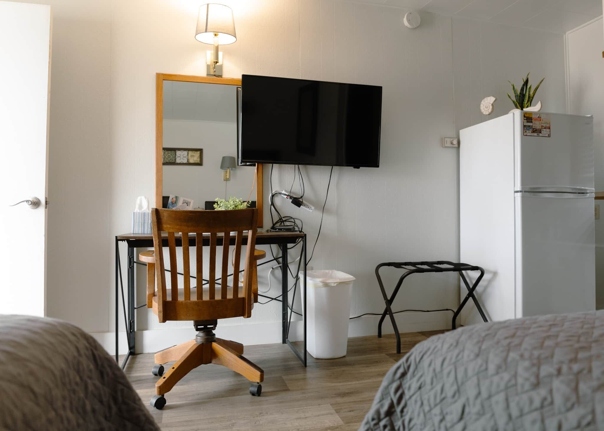 Habitación compacta con una silla de madera, un escritorio, un televisor montado en la pared, una mesita pequeña y un refrigerador.
