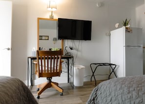 Habitación compacta con una silla de madera, un escritorio, un televisor montado en la pared, una mesita pequeña y un refrigerador.