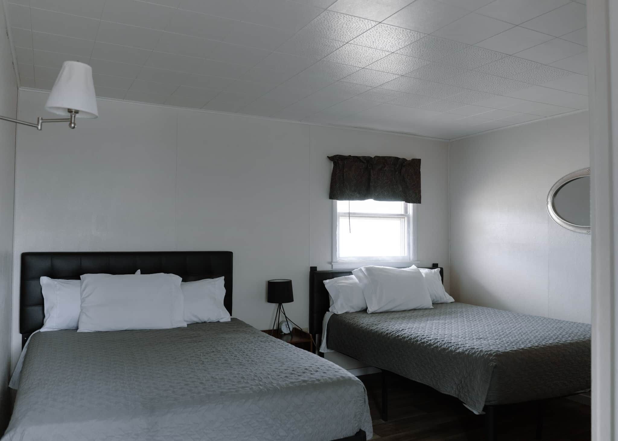 Une chambre avec deux lits, une fenêtre avec un rideau, un miroir rond et une lampe fixée au mur.
