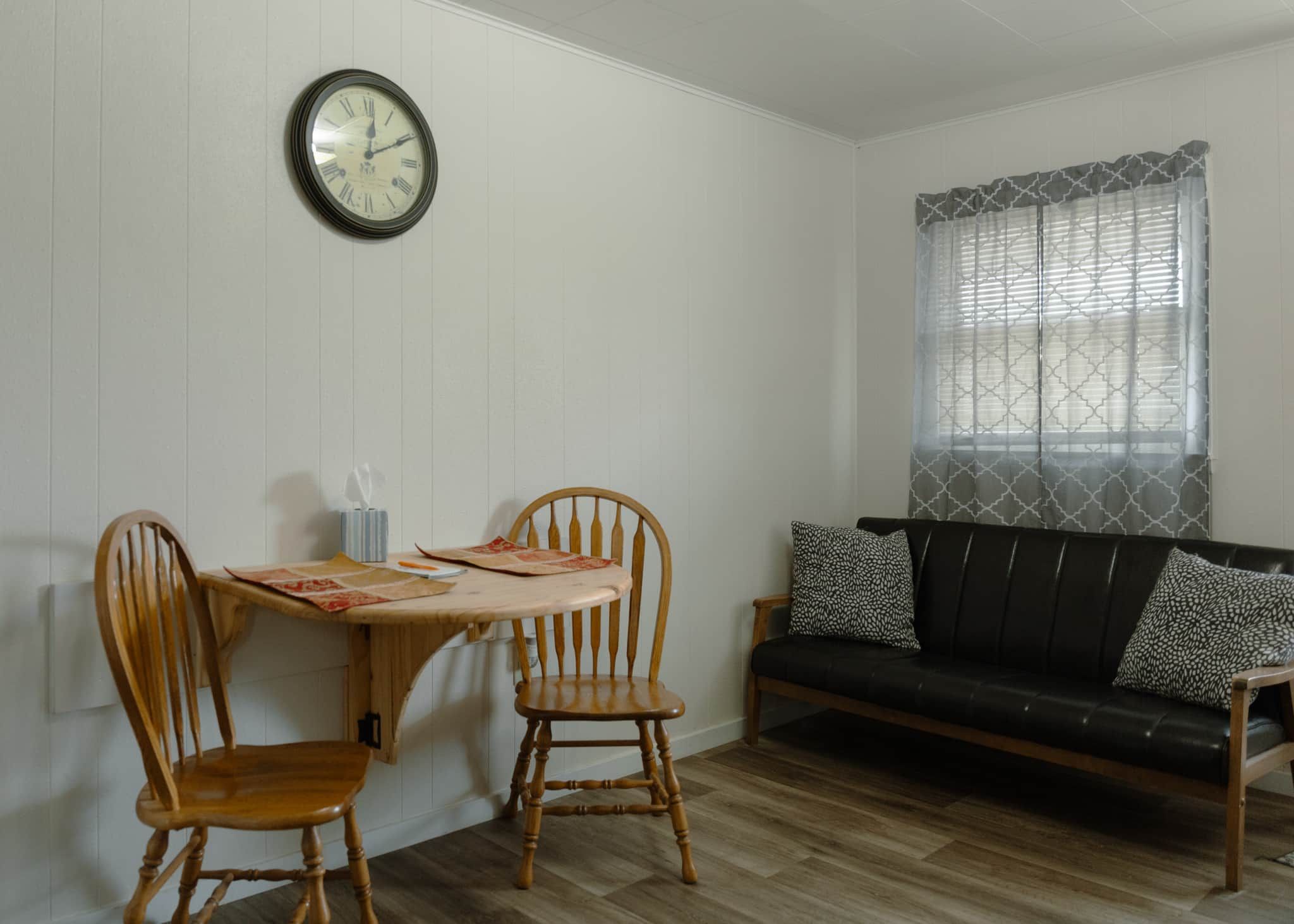 A room with a dining table, two wooden chairs, a black sofa with patterned cushions, and a wall clock.