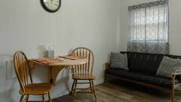 A room with a dining table, two wooden chairs, a black sofa with patterned cushions, and a wall clock.