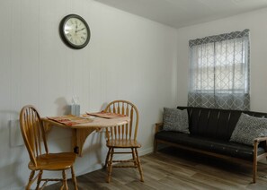 Une pièce comprenant une table à manger, deux chaises en bois, un canapé noir avec des coussins à motifs et une horloge murale.