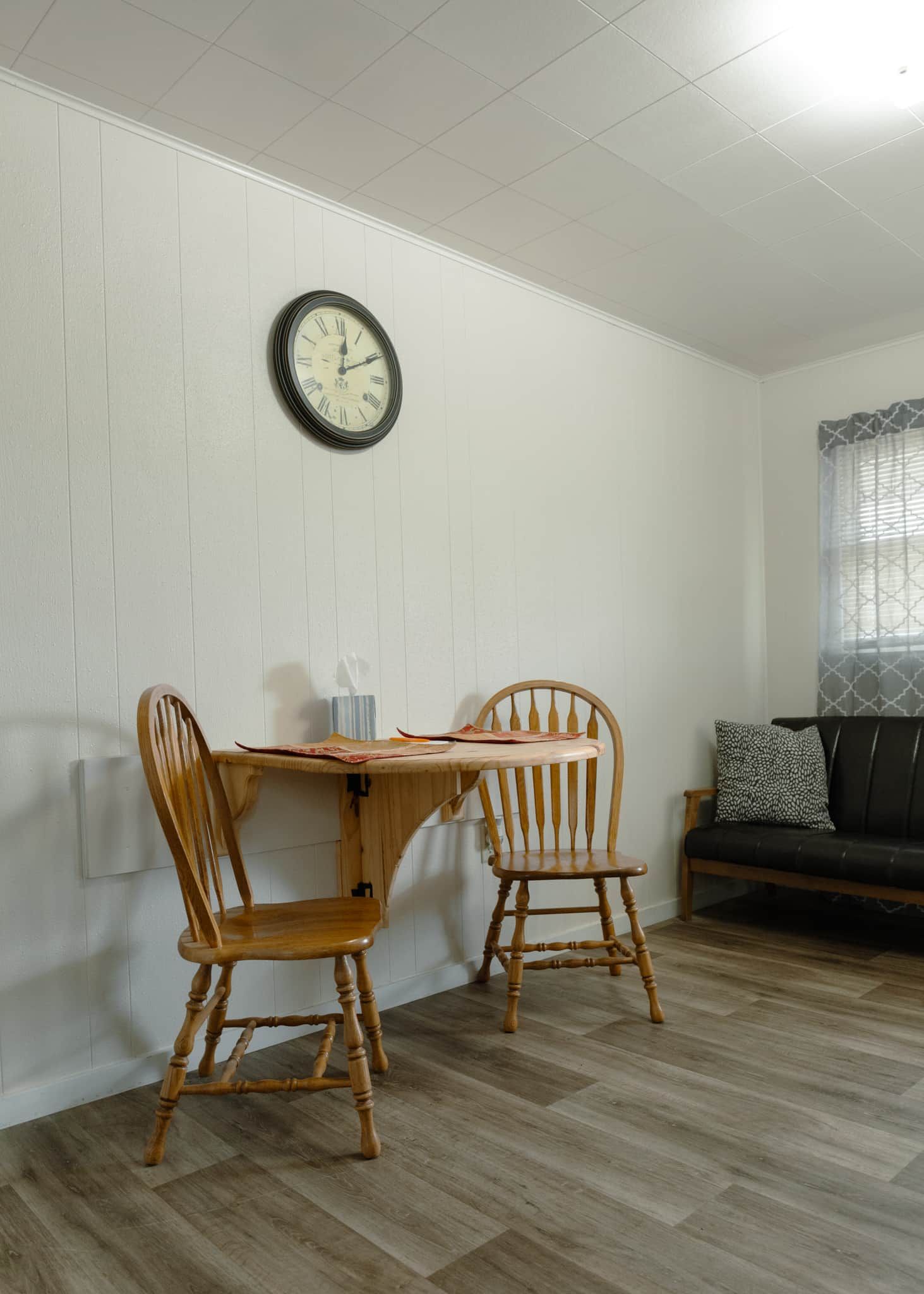 Un comedor con una mesa de madera y dos sillas, un reloj en la pared y una ventana con cortinas estampadas.