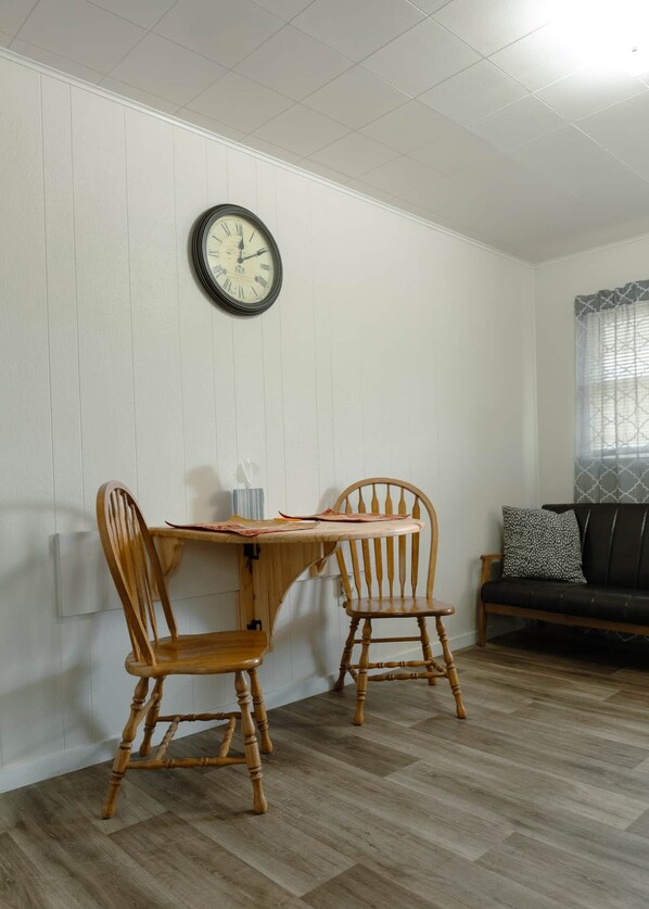 A dining area with a wooden table and two chairs, a wall-mounted clock, and a window with patterned curtains.