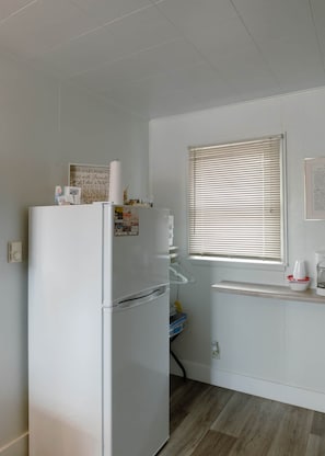 Cozinha compacta com geladeira branca, janela com persianas e bancada com cafeteira.