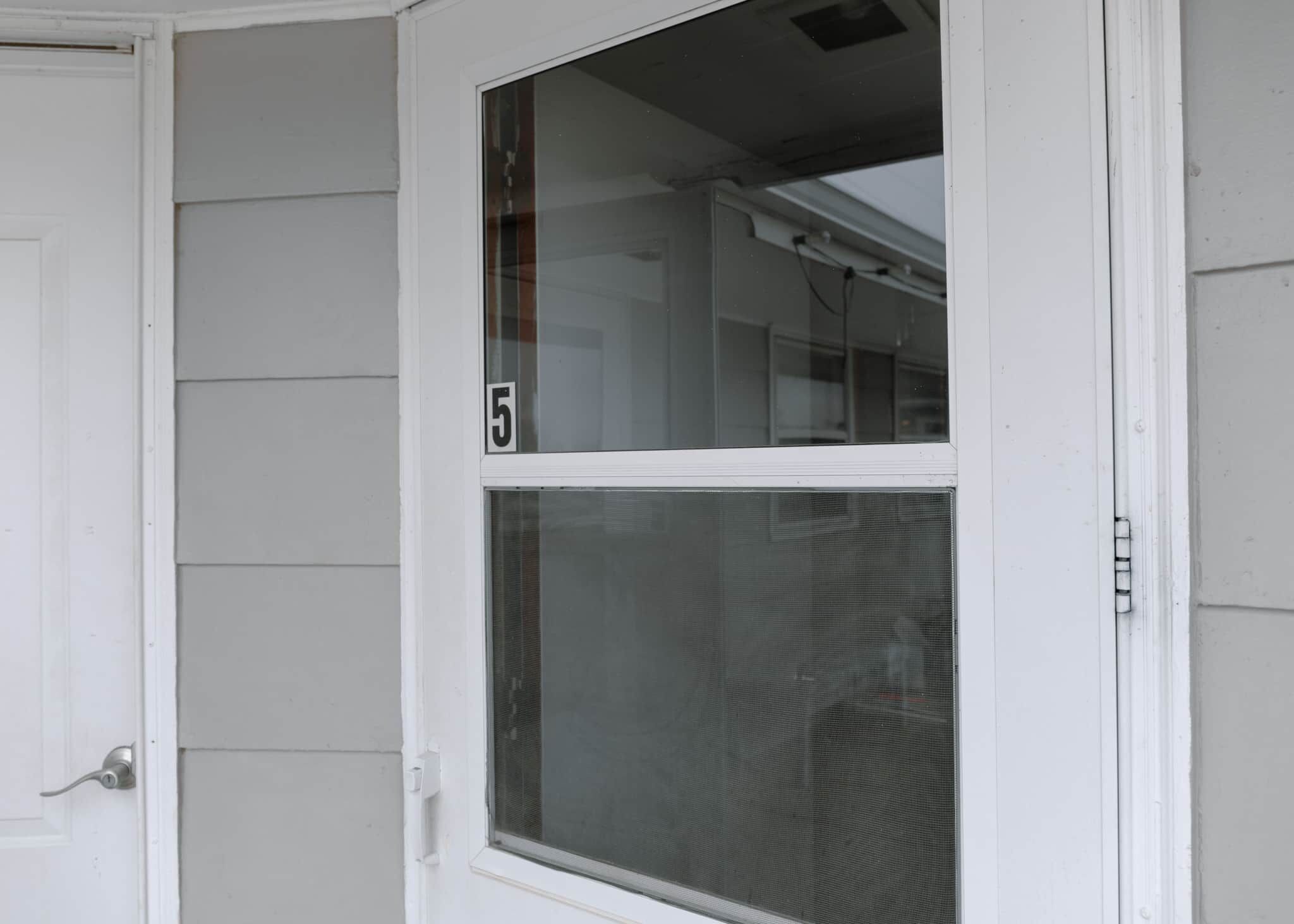 Een witte deur met het cijfer '5', een raam met hor en een witte buitenmuur.