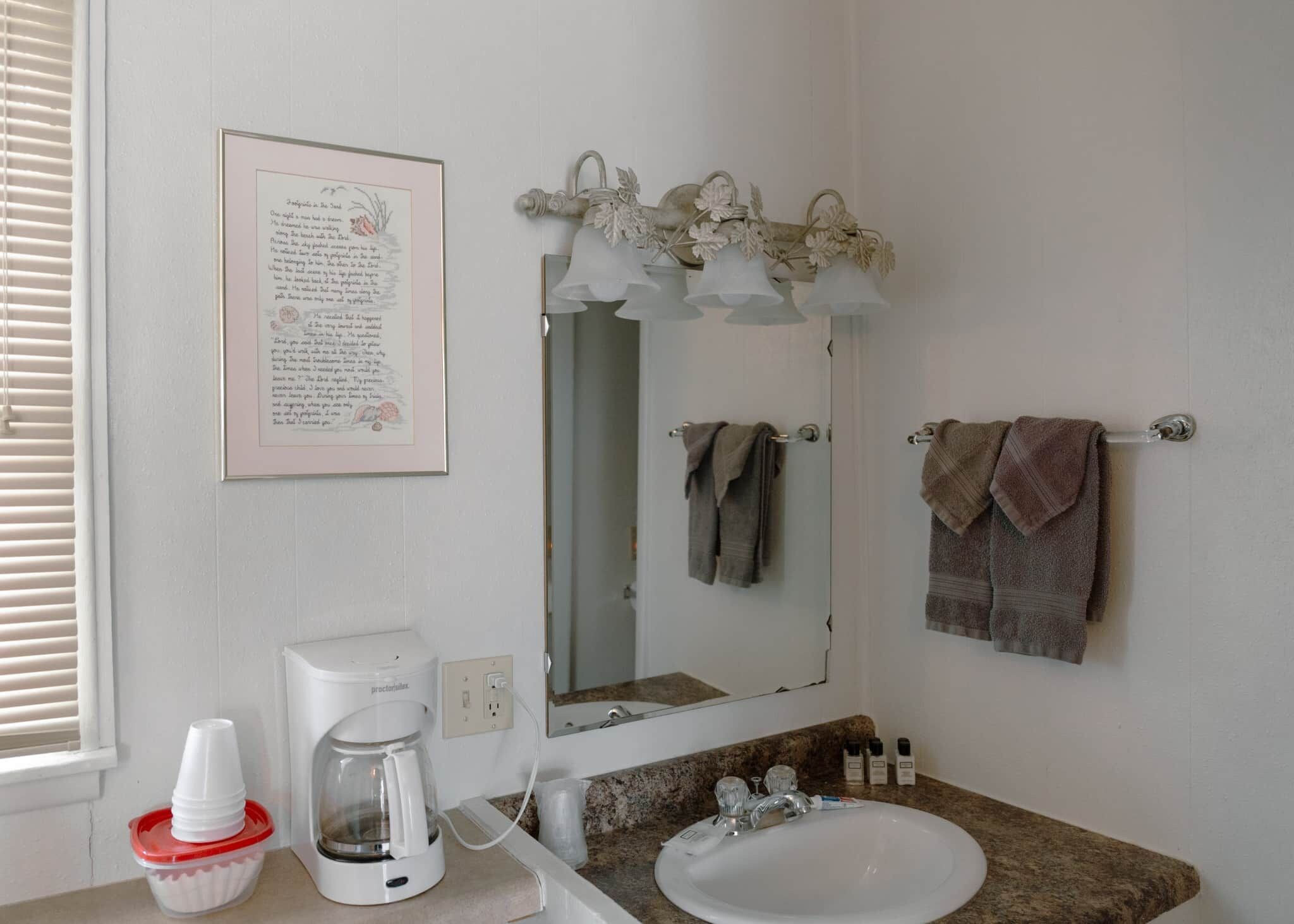 Een badkamer met een wit wastafel, een koffiezetapparaat, een inlijsting gedicht aan de muur, en twee handdoeken die aan haken hangen.