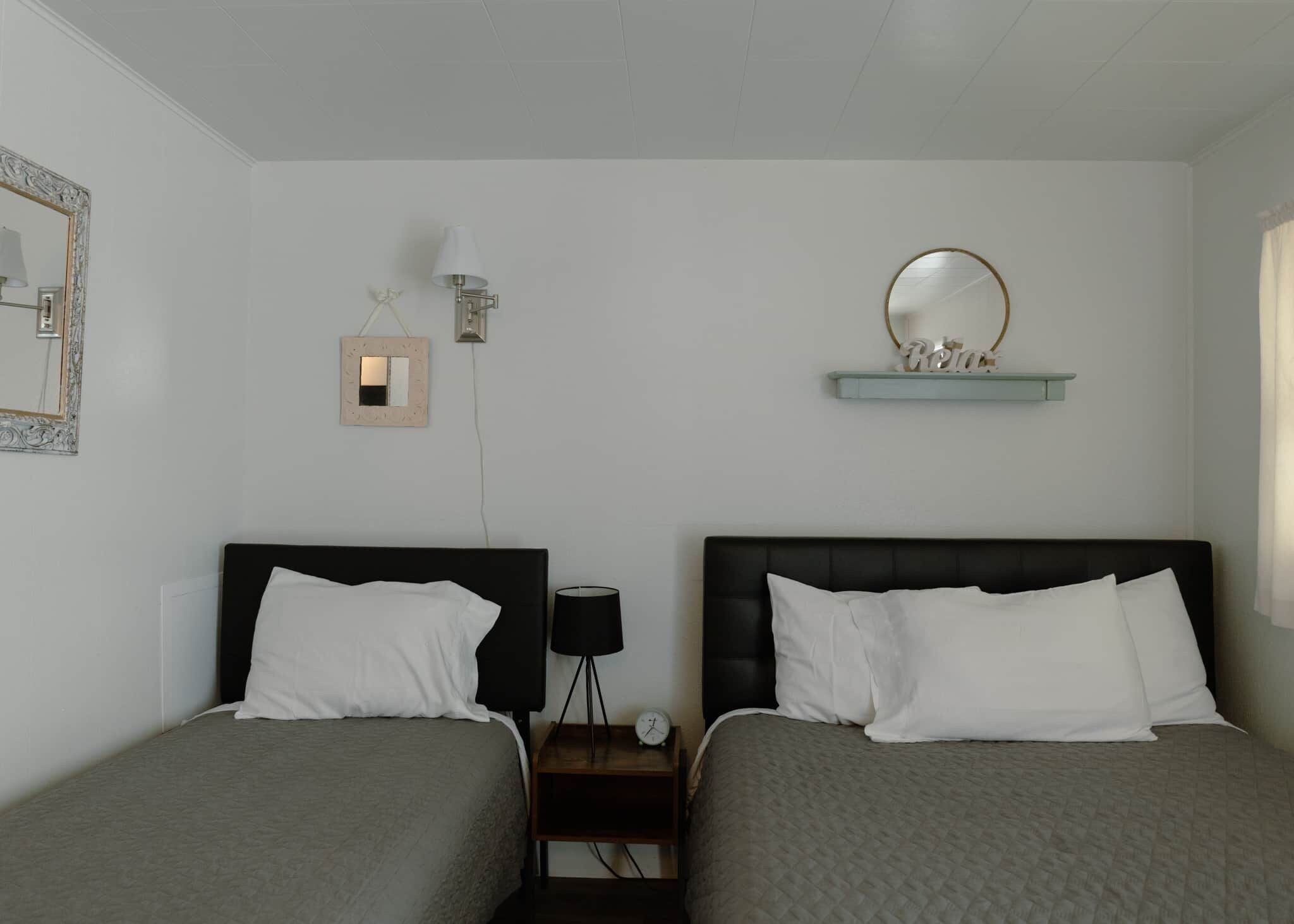 Quarto de hotel com duas camas, mesa de cabeceira com abajur, espelho fixado na parede e prateleira com objetos decorativos.