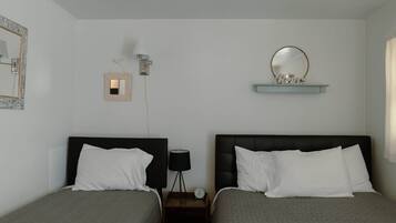 Quarto de hotel com duas camas, mesa de cabeceira com abajur, espelho fixado na parede e prateleira com objetos decorativos.