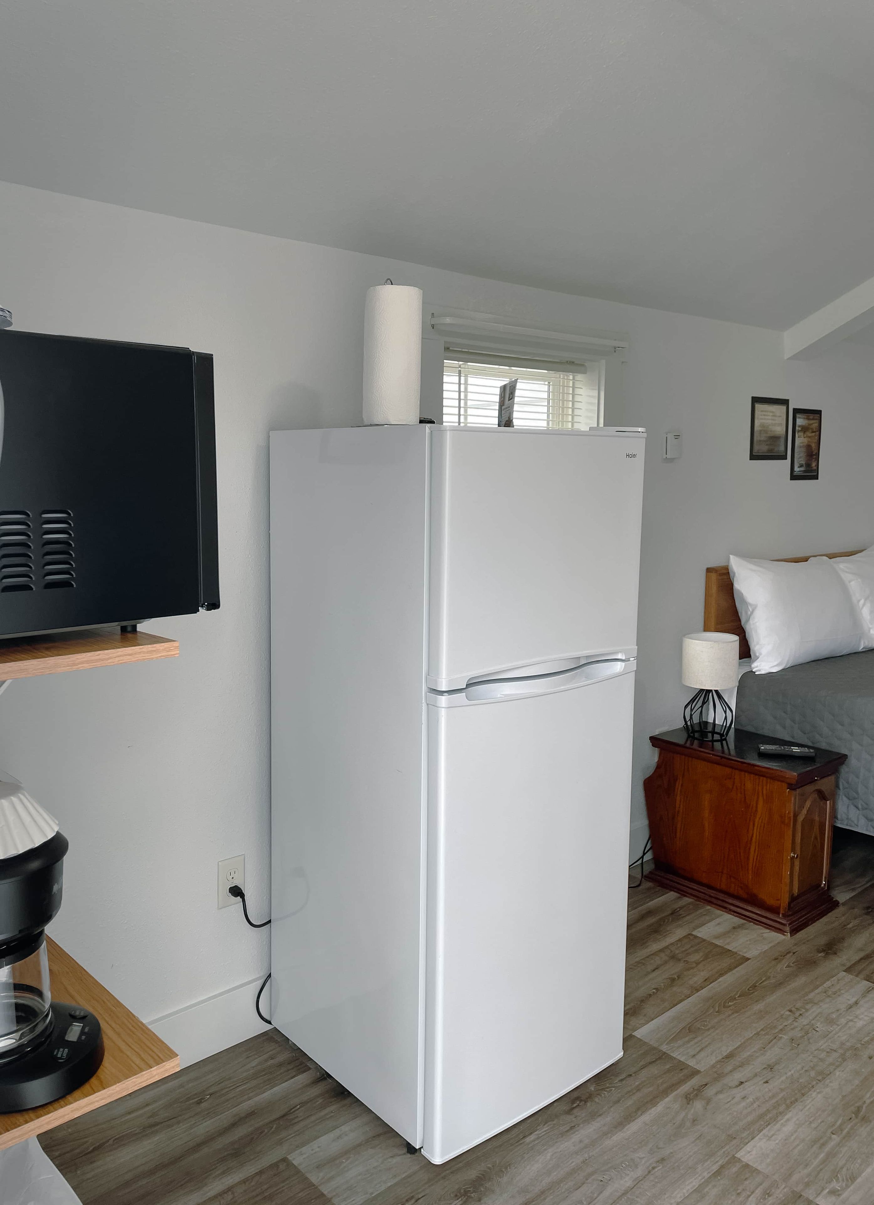 A compact kitchenette with a white refrigerator, a coffee maker, and a wooden shelf.