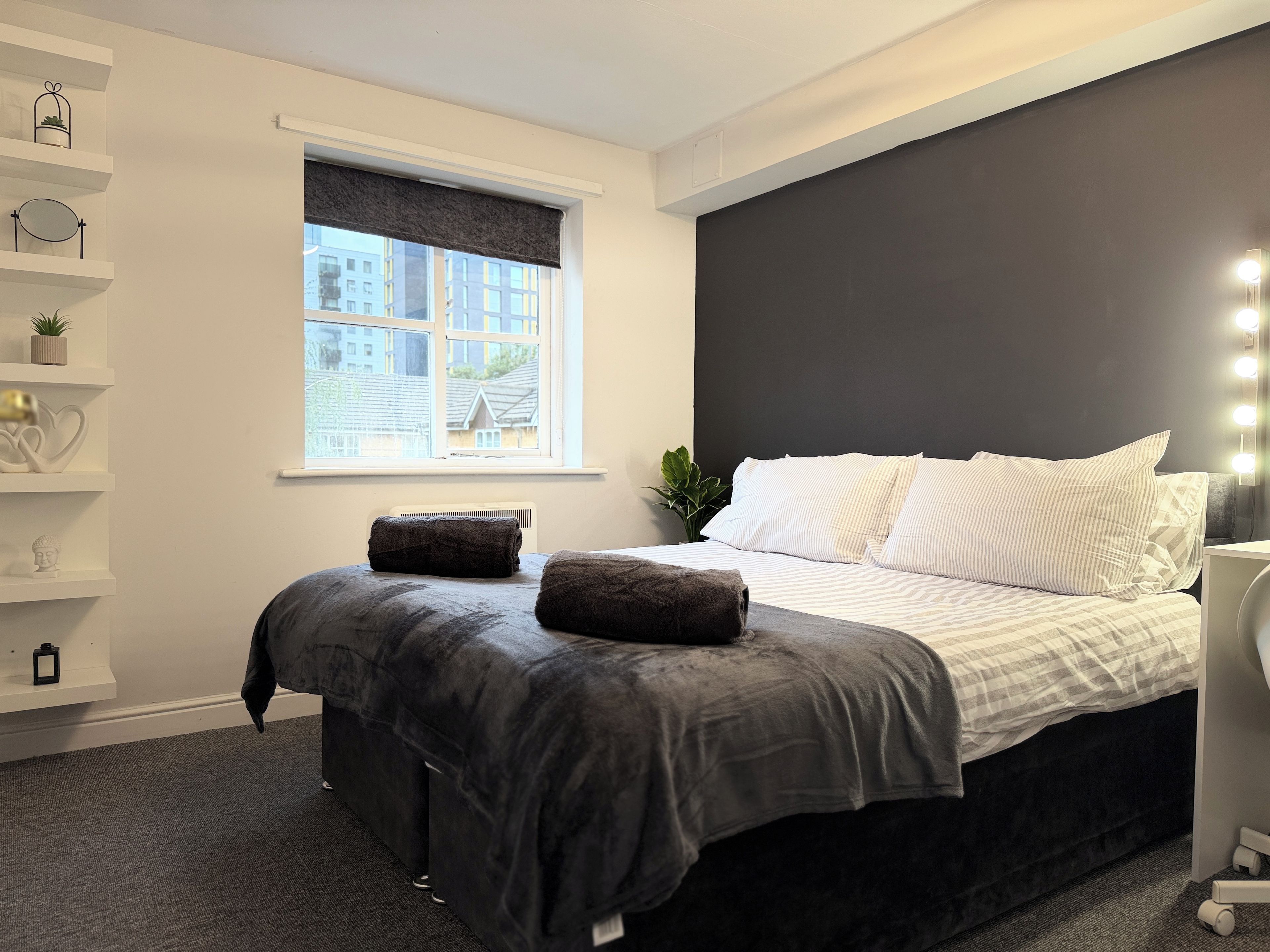 Une chambre moderne avec un lit, des tables de chevet, une fenêtre avec des stores et une vue sur les bâtiments environnants.