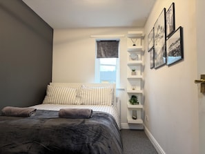 Ein Schlafzimmer mit einem Bett, einem Fenster, einem an der Wand befestigten Regal mit Dekoration und gerahmten Bildern.