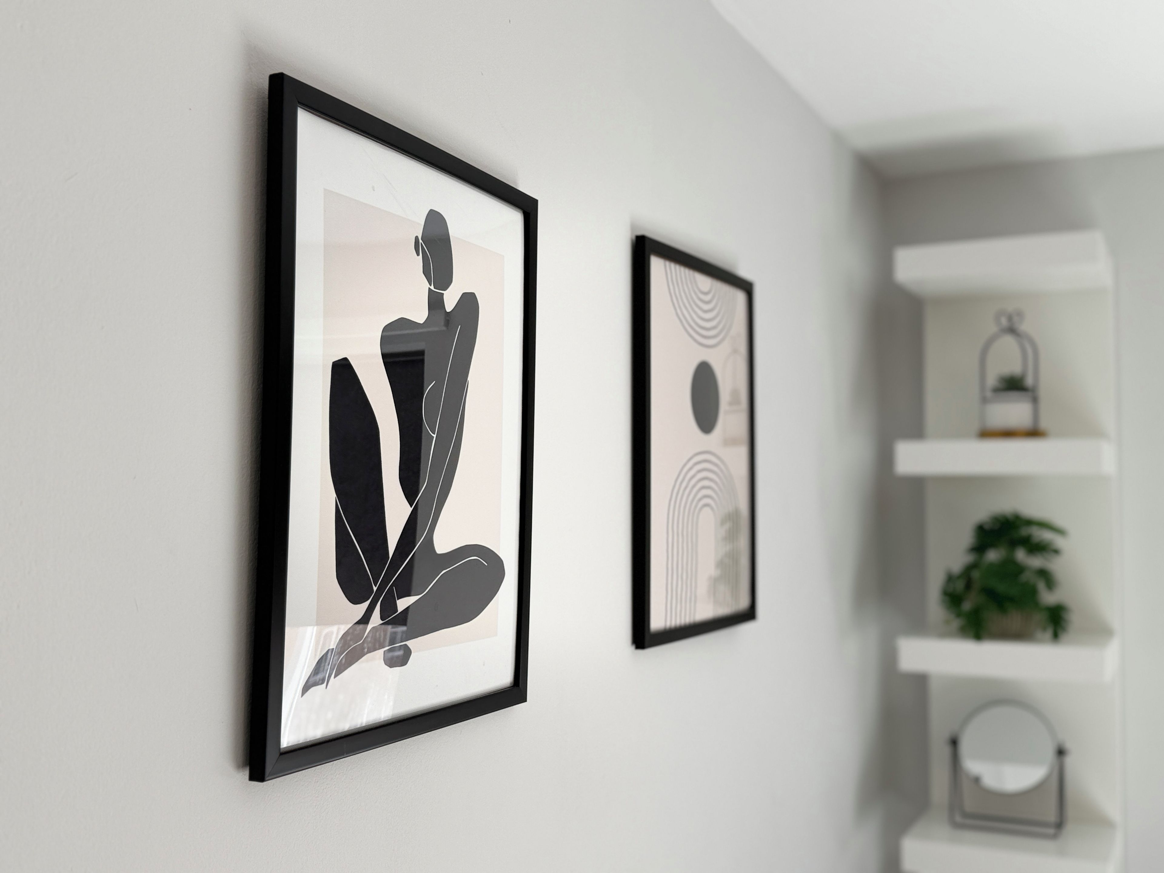 Deux œuvres d’art abstraites encadrées sont accrochées à un mur, avec une étagère blanche et une plante en pot en arrière-plan.