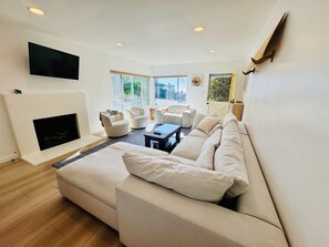 A modern living room with a large sofa, a fireplace, and a flat-screen TV mounted on the wall. - Ocean View 3-brm House w/ Sauna, Fireplace & EV Charging (Hermosa Beach)