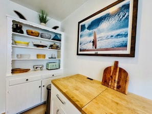 Une cuisine avec des meubles blancs, un plan de travail en bois et une photographie encadrée représentant un surfeur sur une plage.
