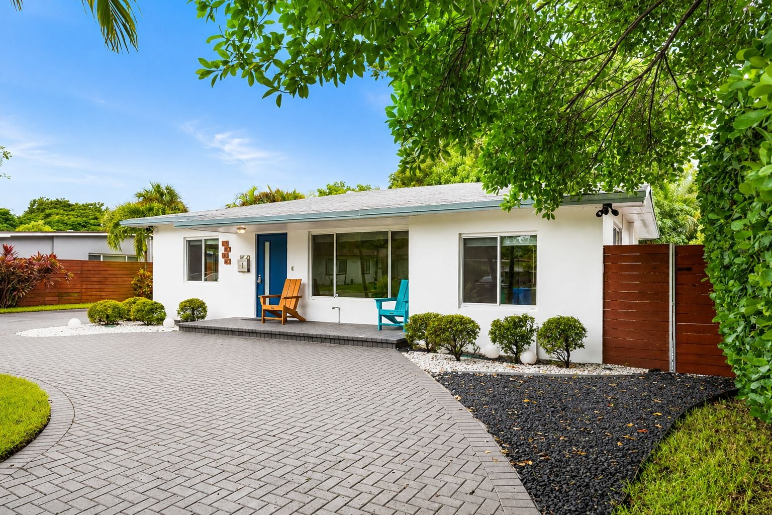 A modern single-story house with a paved driveway, a front porch with two chairs, and a wooden fence.