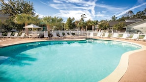 Una piscina limpia con sillas de descanso y sombrillas, rodeada por una valla blanca y árboles.