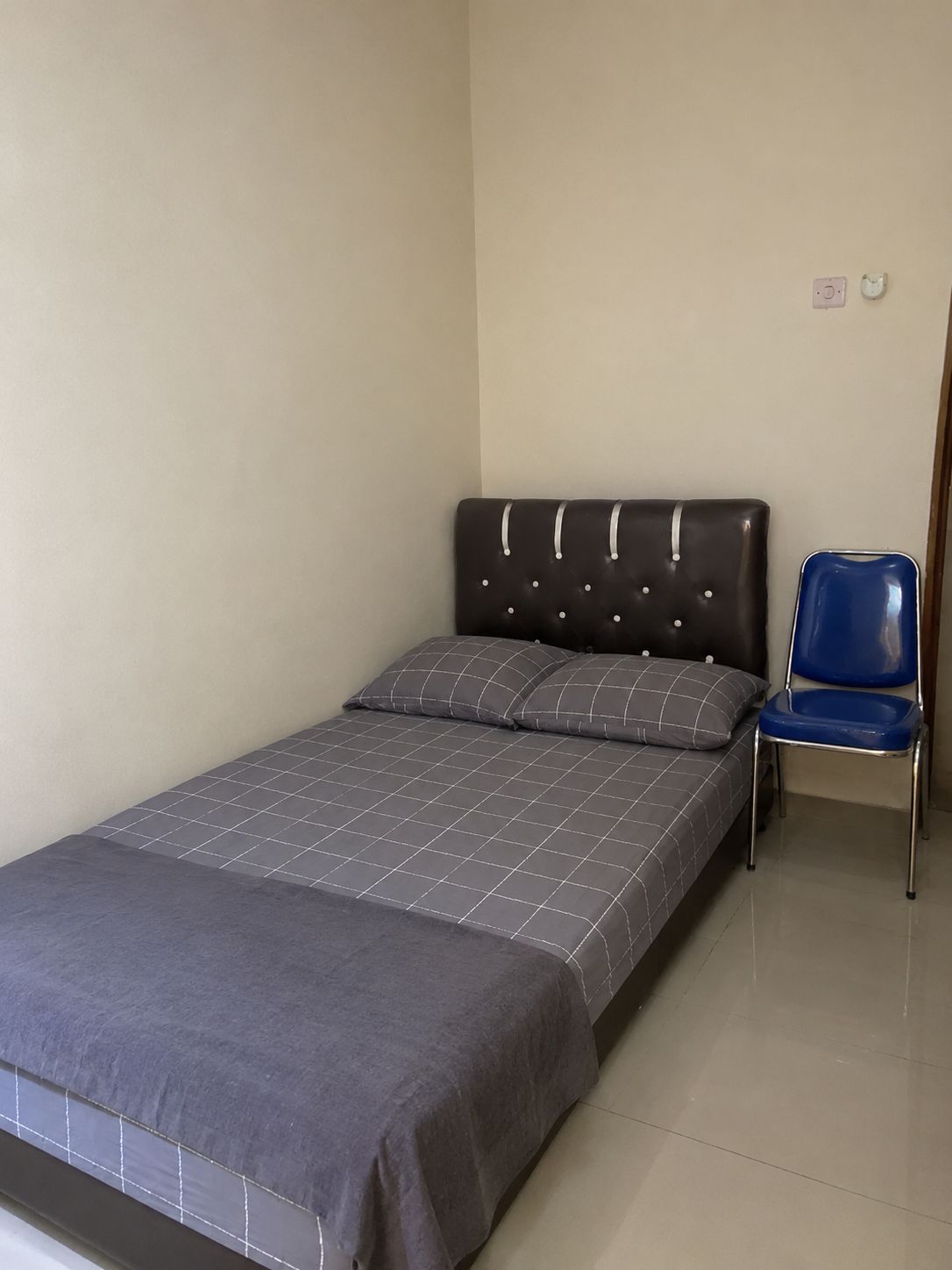 A single bed with a grey bedspread and two pillows, a blue chair, and a plain wall with a light switch.