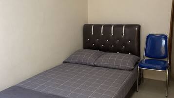 A single bed with a grey bedspread and two pillows, a blue chair, and a plain wall with a light switch.