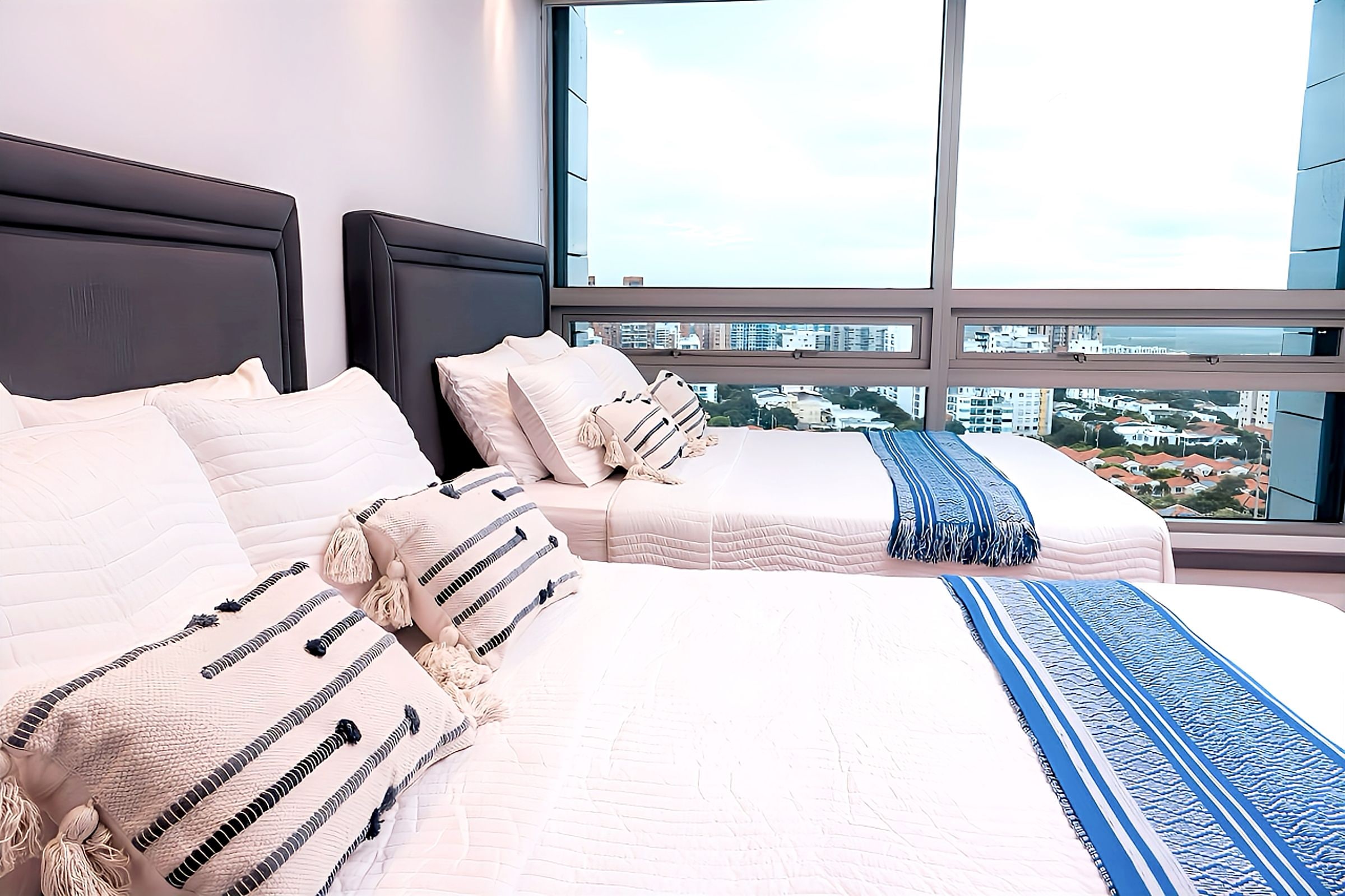 Ein Hotelzimmer mit einem großen Bett, zwei Kissen und Blick auf eine Stadtlandschaft durch große Fenster.