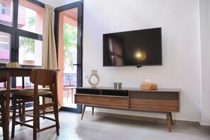 A modern living room with a flat-screen TV mounted on the wall, a wooden entertainment unit, a dining area with a chair, and a view through an open door. - M17—Elegant Duplex in Majorelle—Pool, Balcony, and Parking (Marrakech)