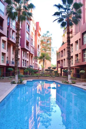 A swimming pool surrounded by multi-story buildings with balconies, palm trees, and a clear sky. - M17—Elegant Duplex in Majorelle—Pool, Balcony, and Parking (Marrakech)