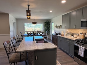 A modern kitchen with a central island, dark cabinetry, and a view of a forest through a large window.