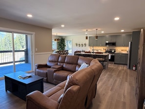 A modern living room with a sectional sofa, a dining area, and a kitchen with grey cabinets and stainless steel appliances.