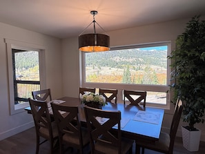 A dining area with a table, chairs, a hanging light fixture, and a large window with a view of a forest and mountains. - Luxury Montana Cabin on Rocky Mountain Front-Entrance to Bob Marshall Wilderness (choteau)