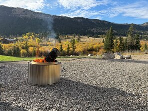A metal fire pit with burning logs in a gravel area, surrounded by a mountainous landscape with autumn foliage.
