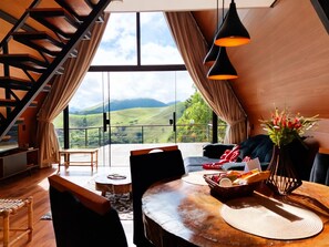 A spacious room with a large window offering a view of rolling hills, a dining area with a table set for two, and a staircase leading to an upper level. - Chalet 4 Elements - Privacy and a breathtaking view 15 minutes from the city center. (Vale do Paquequer)