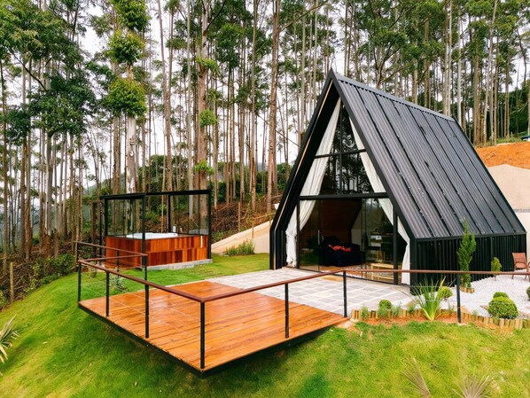 Una cabaña moderna con techo inclinado, una terraza de madera y un ventanal que da a un interior con un sofá rojo.