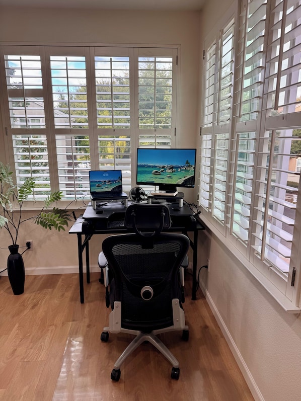 A home office with two monitors, a desk, a chair, and a view through large windows.