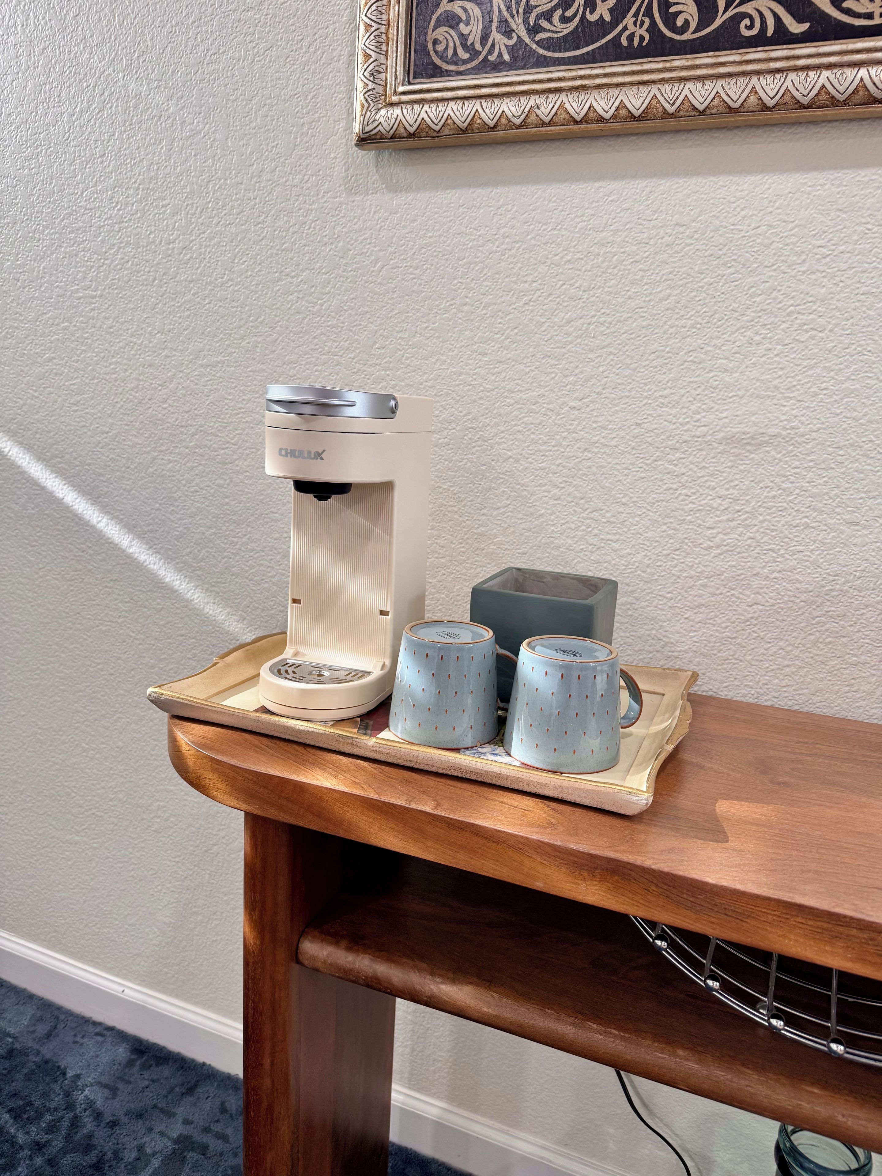 En kaffebryggare står på ett bricka tillsammans med två koppar och en ask, placerat på ett träbord framför en vägg med en tydlig struktur.
