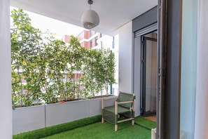 Un balcón con piso de césped artificial verde, una silla de mimbre y vistas a edificios a través de un muro blanco.