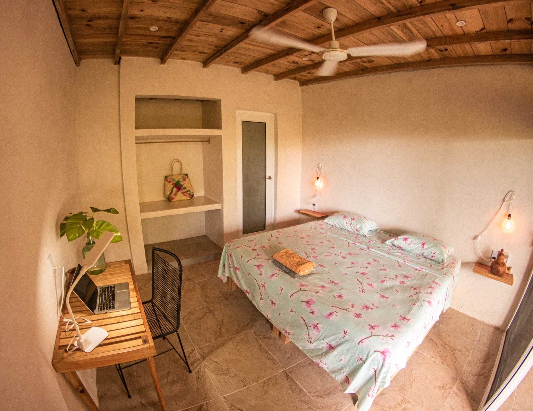 Un dormitorio con una cama, un escritorio con una computadora portátil, una silla, un ventilador de techo y una estantería con una bolsa.