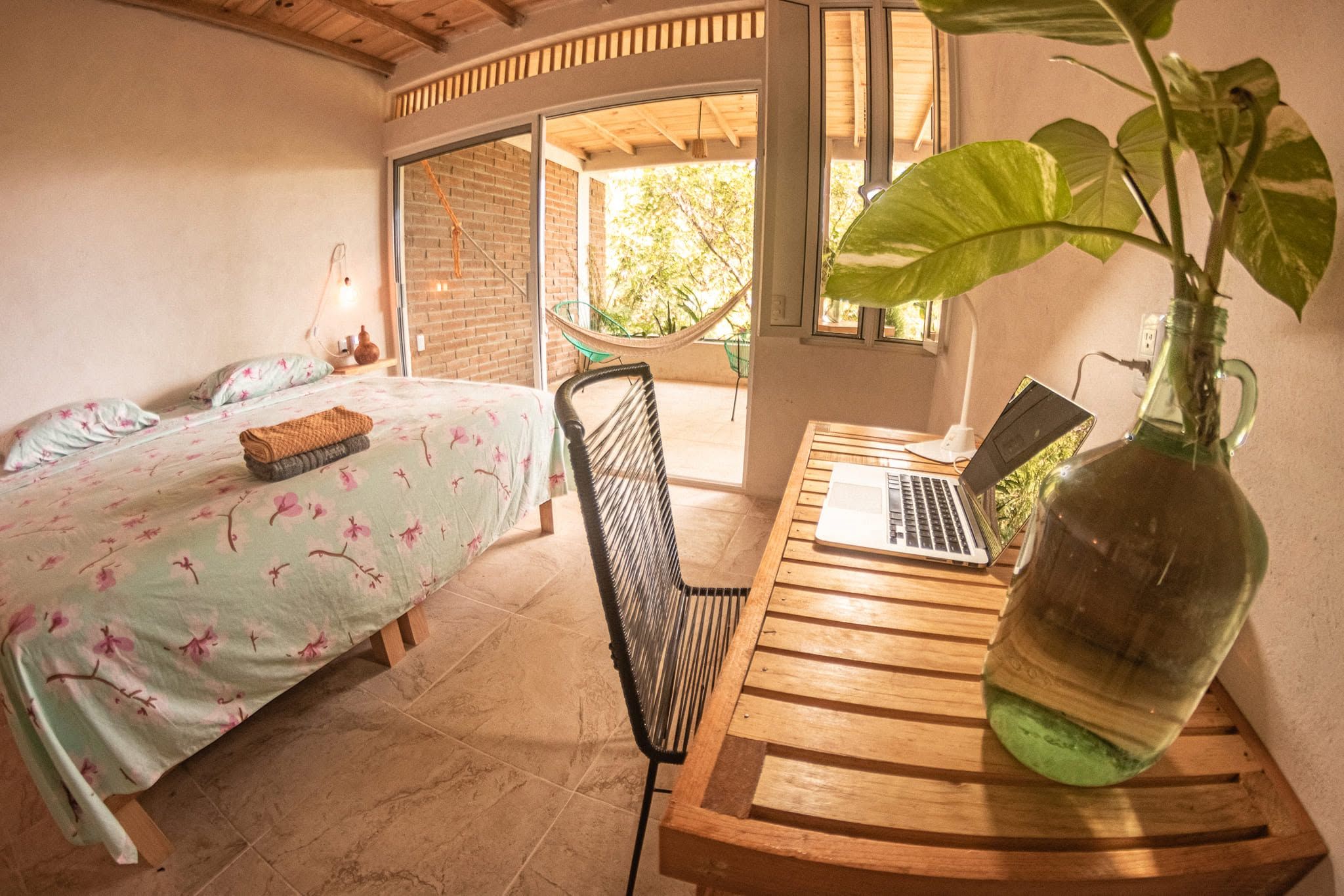 Ein Schlafzimmer mit einem Bett, einem Laptop auf einem Holztisch und einer großen Pflanze in einer Flasche.