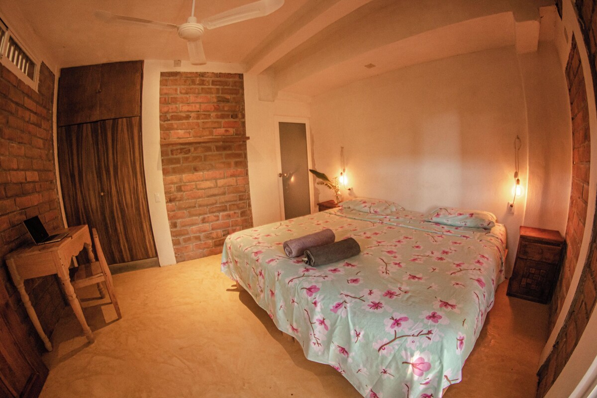 A bedroom with a brick wall, a wooden door, a bed with floral bedding, a wooden nightstand, and a ceiling fan.