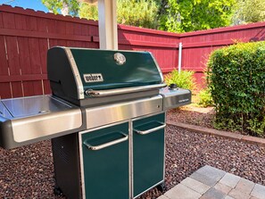 A Weber Genesis portable grill with a stainless steel finish, a side burner, and a storage drawer, set against a red wooden fence and greenery.