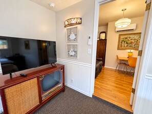 A living room with a flat-screen TV on a wooden entertainment unit, a dining area with a table and chairs, and a hallway leading to a bedroom.