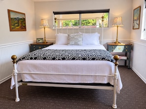 A neatly made bed with a patterned bedspread, two bedside tables with lamps, and a window with a view of greenery.