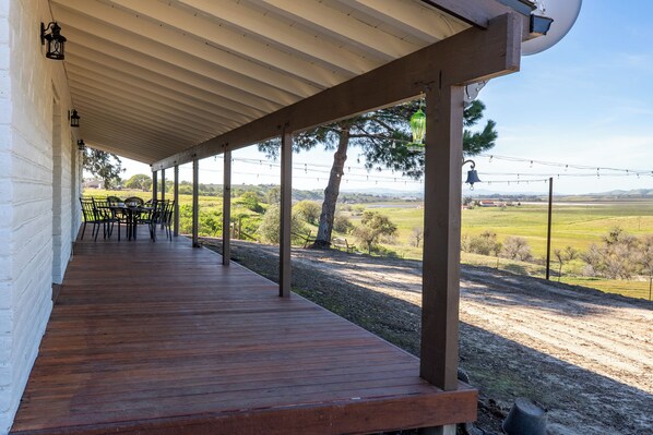 Un espacio al aire libre techado con una terraza de madera, mesas y sillas, con vistas a un paisaje rural.