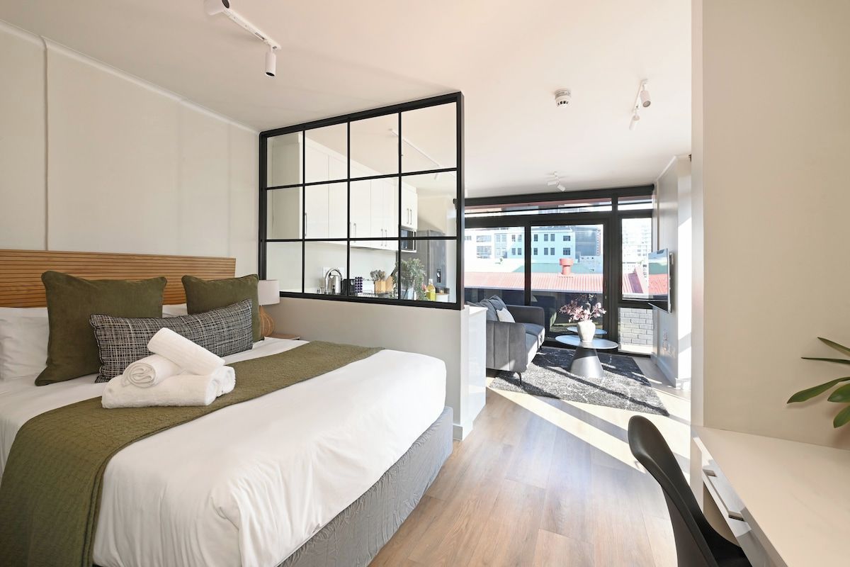 A modern hotel room with a large bed, a dining table, and a view of the city through a large window.