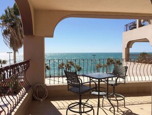 Varanda com mesa e cadeiras, vista para a costa, palmeiras e o mar.
